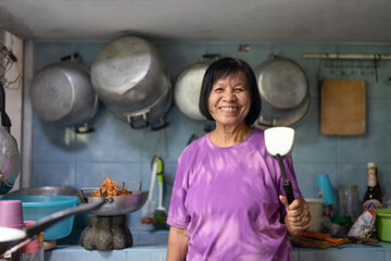 Portrait of Asian elderly woman holding stainless steel turner while cooking Thai food. Smiling grandmother preparing food in Thai kitchen room for family dinner - obrazy, fototapety, plakaty