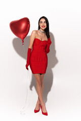 Full length of stylish model in red dress holding heart shaped balloon on white background with shadow