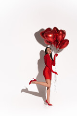 Full length of pretty woman in red heels and dress posing with heart shaped balloons on white background with shadow