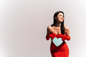 Cheerful woman in red dress with naked shoulders holding paper heart isolated on grey