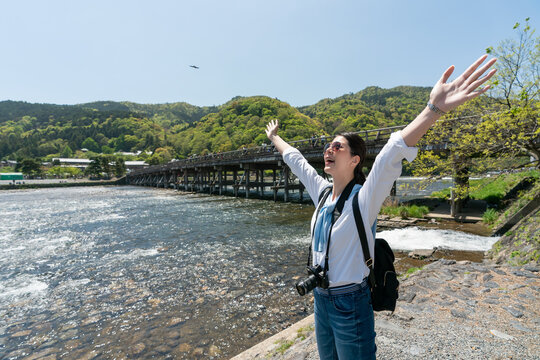 happy asian Japanese woman visitor standing Katsura river with opening arms while enjoying fresh air and natural beauty with Togetsu-kyo Bridge at background in arashiyama Kyoto japan