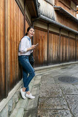 full length of cheerful asian Japanese woman visitor learning about Gion Ishibeikoji lane on phone while traveling to Kyoto japan. she leans against wooden wall and admiring the historic alley