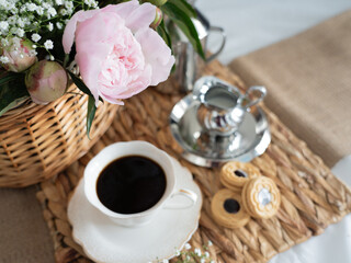 An artistic still life with pink peonies in a wicker basket, a cup of coffee, cream and cookies. Top view. Horizontal photo.