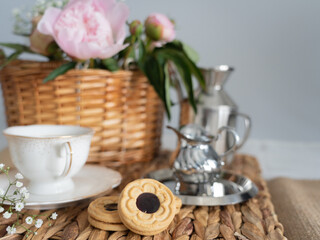 An artistic still life with pink peonies in a wicker basket, a cup of coffee, cream and cookies. Horizontal photo.