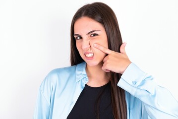 Young caucasian woman wearing blue overshirt over white background pointing unhappy to pimple on forehead, ugly infection of blackhead. Acne and skin problem