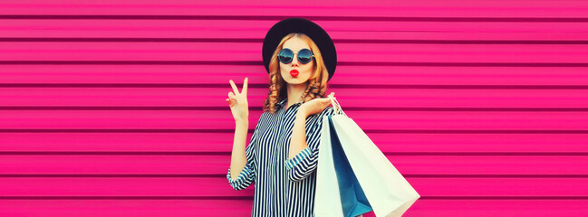 Portrait of beautiful young woman with shopping bags blowing her lips sends air kiss wearing striped shirt, black round hat on pink background