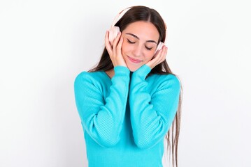 Fototapeta premium Pleased Young caucasian woman wearing blue sweater over white background enjoys listening pleasant melody keeps hands on stereo headphones closes eyes. Spending free time with music