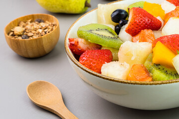 Fresh fruit salad in a bowl. Multicolored and tropical fruits. Pineapple, mango, grape, strawberry, papaya, melon, kiwi. Additional with chestnuts and granola. Selective focus
