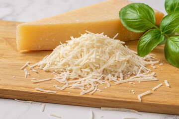 Shredded grana padano cheese on a cutting board. Italian parmesan cheese whole wedge and grated with green basil herb over wooden background. Delicious hard cheese. Dairy product. - obrazy, fototapety, plakaty