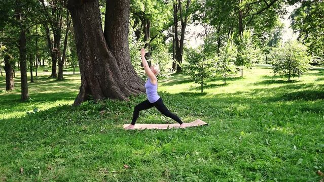 Middle aged caucasian woman in sportswear doing yoga outdoors in warrior pose, Virabhadrasana I. The concept of yoga, stretching, healthy lifestyle. Sports activities in nature. 4k footage