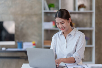 Beautiful young Asian businesswoman sitting working with laptop thinking about new ideas for her work with a bright and happy face.