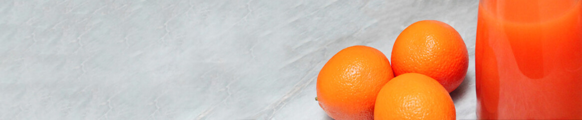 part of a glass with bright orange juice and three whole orange fruits on the right side of the image on a light gray uneven background with space for text