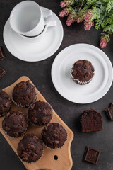 On a dark table are chocolate muffins on a cutting board, tea cups and flowers.