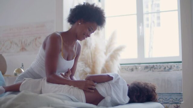 Young mother playing with her daughter in bed after waking up. Happy Black woman and cute little girl tickling each other, fighting and laughing, having fun together on weekend. Happy family concept.