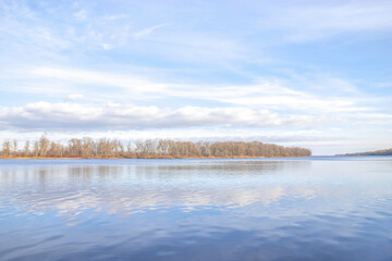 View across wide calm river in late fall with treed island in distance, cloudy sky reflected in water, daytime, nobody