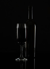 A tall glass of red wine and a bottle on a black table. White highlights on the glass and bottle. Black background