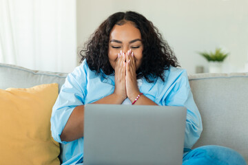Great news concept. Emotional ecstatic black woman celebrating online win sitting on sofa and using laptop, copy space