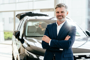 Portrait of positive smiling happy man buyer in dealership, stand happily posing next to new car, wearing formal suit. Male agent standing crossed arms in car showroom.