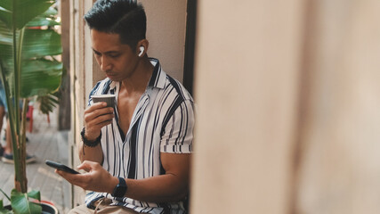 Young man sits outside drinking coffee and uses mobile phone