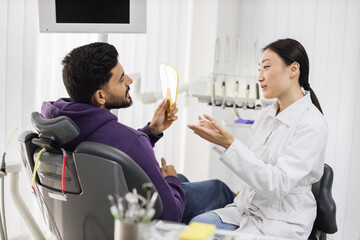 Portrait of handsome bearded male checking his teeth while looking at mirror after curing teeth while smiling beautiful female asian dentist looking at patient in new bright dental clinic.