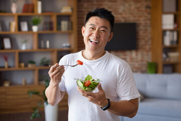 Happy asian mature man holding fork and bowl with fresh vegetable salad, eating healthy lunch after domestic training - obrazy, fototapety, plakaty