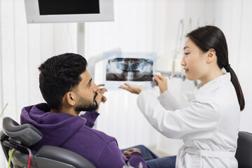 Back view of young attractive man visiting dentist, sitting in dental chair at modern light hospital clinic. Young asian woman dentist holding x ray image. Focus on picture.