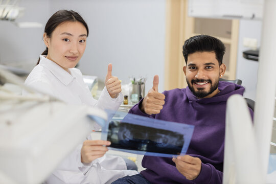 Attractive young bearded man, sitting on dentist chair and looking at x-ray picture of scan image of teeth together with his cheerful female asian dentist at clinic showing thumb up.
