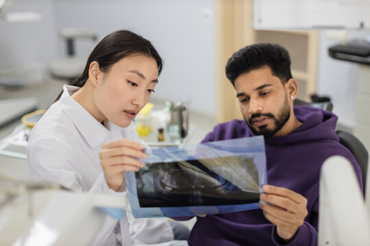 Focus on x ray picture, attractive young bearded man, sitting on dentist chair and looking at x-ray scan image of teeth together with his cheerful female asian dentist at clinic.