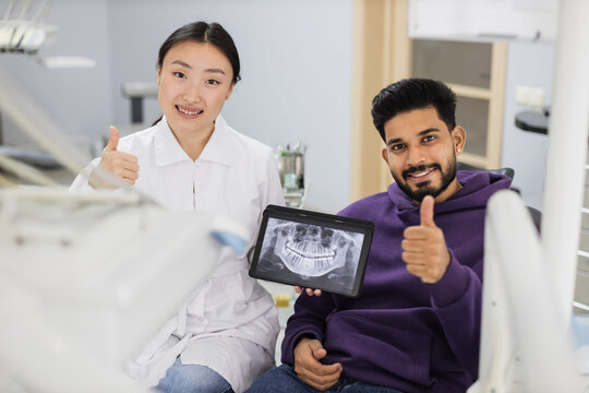 Smiling happy bearded man visiting dentist, sitting in dental chair at modern light hospital clinic. Young asian woman dentist holding tablet pc with x ray scan showing thumbs up.