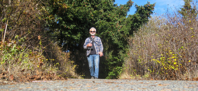 Senior man walking along a footpath with binoculars and a camera, Saanich, Vancouver Island, British Columbia, Canada