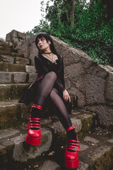 Young and skinny hispanic goth girl with black dress, pantyhose and red platforms shoes seated in a stone stairs next to white flowers in the forest