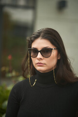 A gorgeous brunette woman wears sunglasses with sunglasses chains. Confident girl in urban portrait.