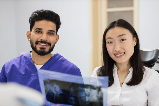 Smiling Happy Asian Woman Visiting Dentist, Sitting In Dental Chair At Modern Light Hospital Clinic. Young Bearded Man Dentist Holding X Ray Image Scan.