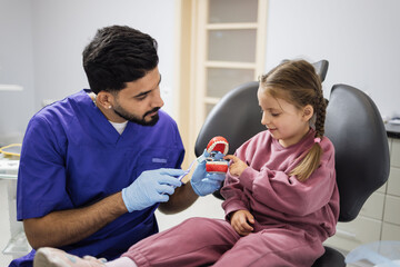 Side view of young confident bearded pediatric dentist showing to little preschool girl how to brush teeth on artificial jaw at light modern dental office. Pediatric dentistry.