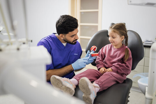 Side view of young confident bearded pediatric dentist showing to little preschool girl how to brush teeth on artificial jaw at light modern dental office. Pediatric dentistry.