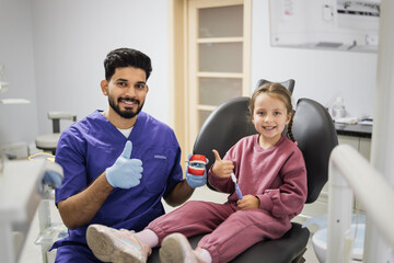 Happy confident male dentist tells little child girl how to brush the teeth on artificial jaw model showing thumbs up. Caries prevention, pediatric dentistry, milk teeth hygiene concept.