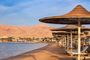 Hotel beach with rows of sun loungers under straw umbrellas against mountains, Dahab, Egypt