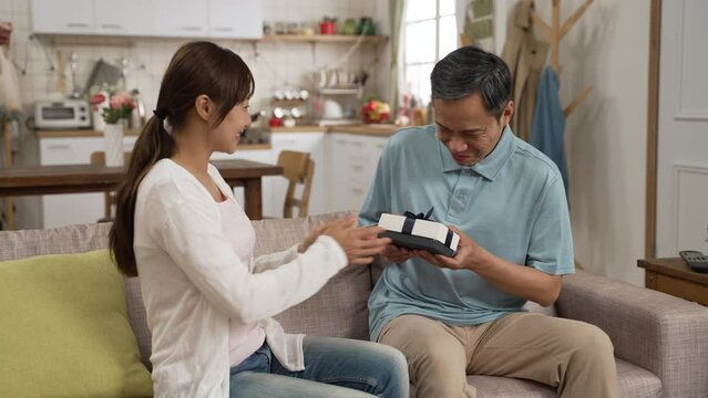 asian adult granddaughter covering grandpa’s eyes and showing him a gift from behind on Father’s Day at home. surprised elderly man saying thanks to her sweet act