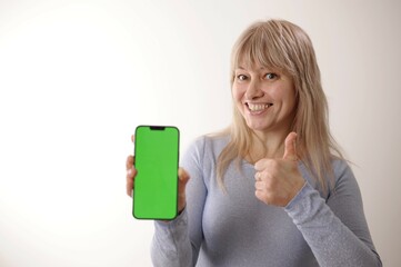 An adult woman 45-50 with blond hair in a blue dress is standing with a mobile phone Green screen chroma key She smiles advertising any products supply demand