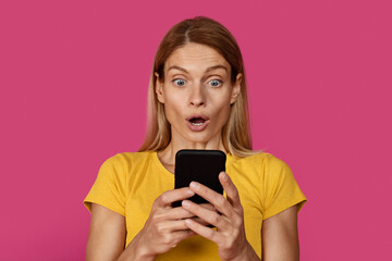 Fototapeta premium Glad surprised mature caucasian blonde female in yellow t-shirt with open mouth looks at smartphone