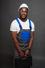Modest engineer in protective helmet and transparent glasses stands against grey wall, holding suitcase with tools in his hand. An endearing black man with box of tools in his hands looks into camera