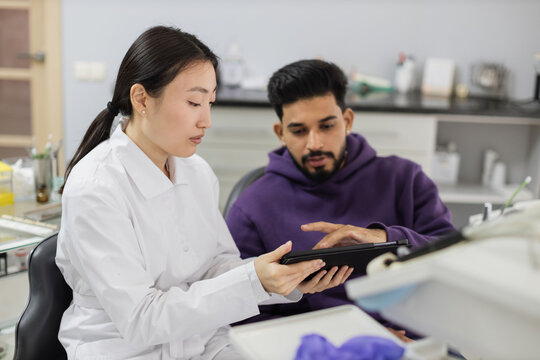 Medicine, pediatric dentistry and oral care concept. Female confident asian dentist showing tablet pc computer to worried male patient, bearded young man at modern dental clinic.
