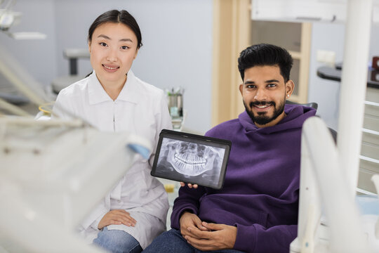 Smiling happy bearded man visiting dentist, sitting in dental chair at modern light hospital clinic. Young asian woman dentist holding tablet pc with x ray scan.