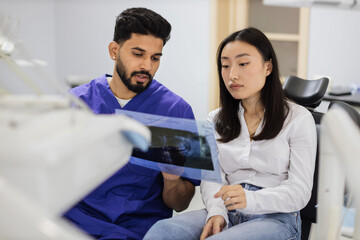 Medicine, dentistry and oral care concept. Male confident dentist showing x ray picture to attractive young patient, asian woman at modern light dental clinic.