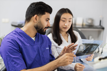 Attractive young asian woman, sitting on dentist chair and looking at x-ray picture of scan image of teeth together with her confident professional male dentist at clinic.