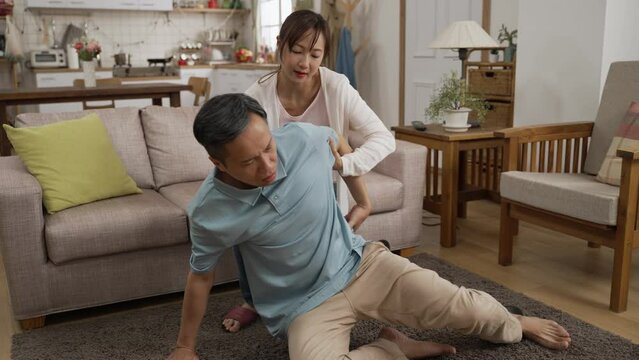 Caring Asian adult granddaughter helping her grandfather sit and rest on sofa after a fall on floor at home. She massages his lower back to make him feel better