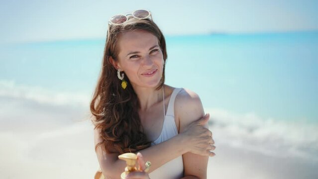 Portrait of smiling stylish 40 years old woman at the beach in white beachwear applying spf.