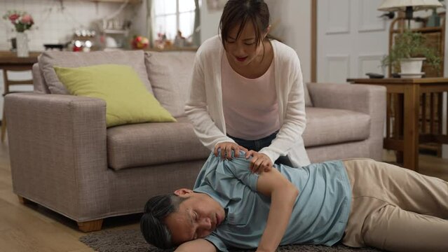 Closeup of anxious Japanese adult daughter calling to her fainted dad lying on floor in coma at home. She tries to wake him up by shaking and slapping him gently