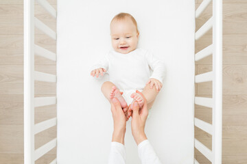 Young mother hands holding and massaging infant cute small bare feet in white crib at home room....