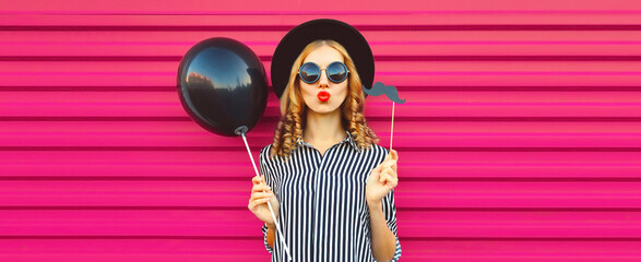 Portrait of funny cheerful young woman having fun holding black balloon, showing mustache on stick...